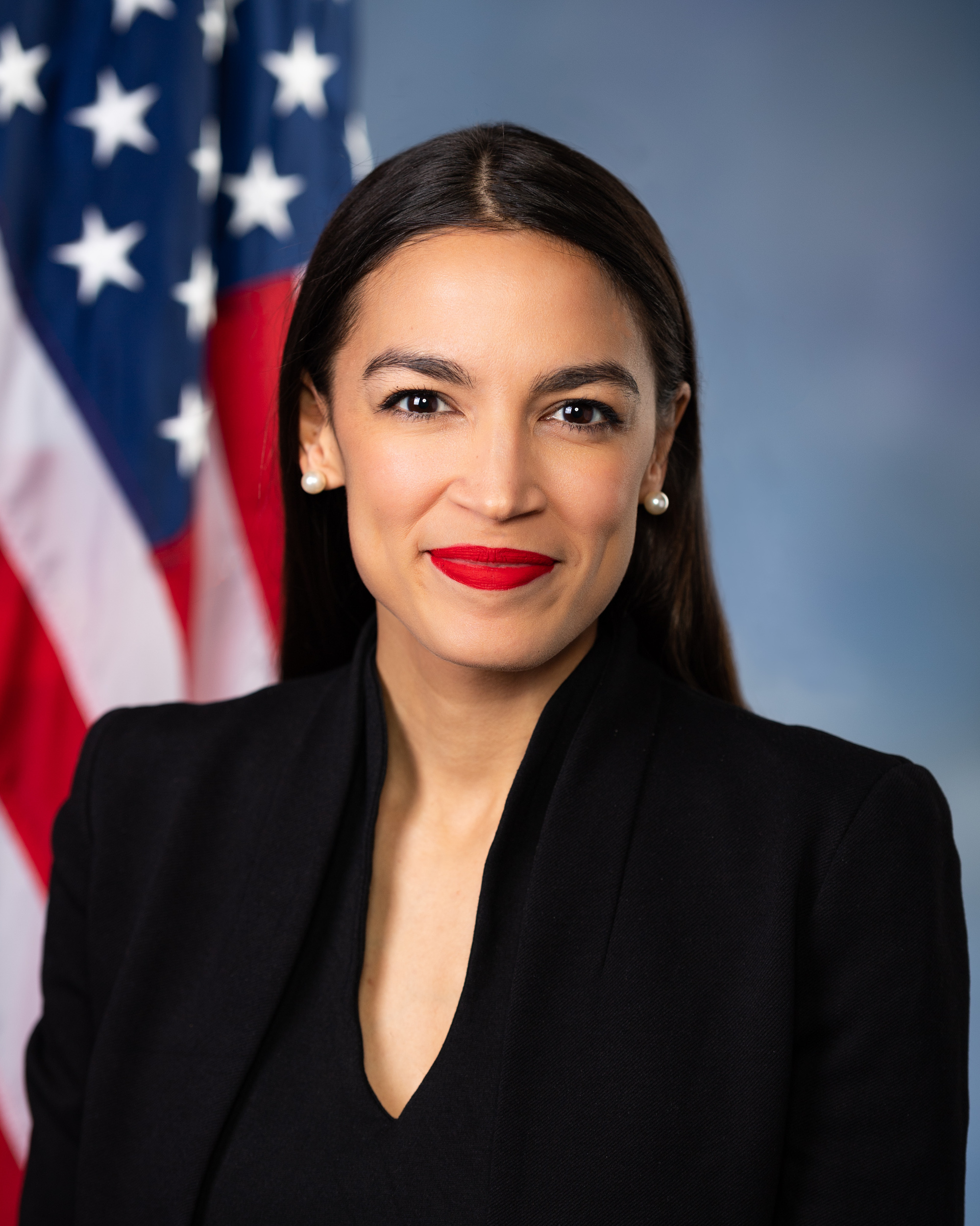 Speaker of the U.S House of Representatives Alexandria Ocasio-Cortez's headshot standing in a black suit with the US flag behind her.